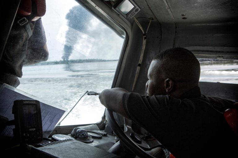 The captain of a small gunboat navigates down the Bonny River as smoke billows from a illegal refinery on fire, Rivers State, Nigeria